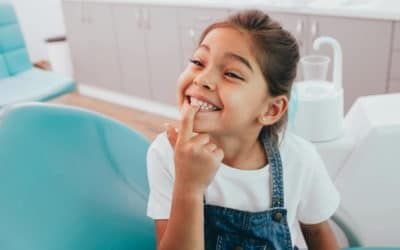 Kleines Mädchen sitzt auf einem Behandlungsstuhl und deutet mit ihrem Finger auf ihre verdrehten Zähne